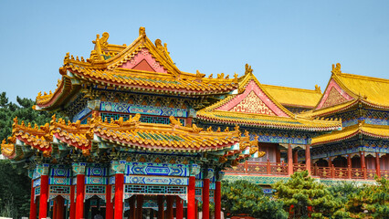 Fototapeta premium Traditional Chinese temple in Penglai, Shandong, with red pillars, golden roofs, and ornate details, surrounded by lush greenery and a serene natural setting.