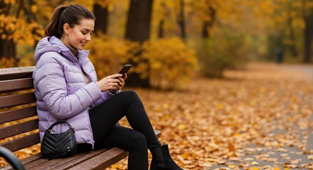A smiling woman uses her smartphone while sitting on a bench in an autumn park. Young person relaxing outdoors with mobile technology. Copy space.