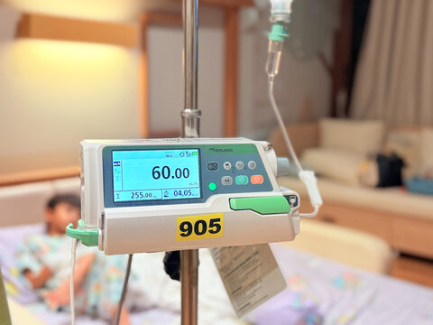 Medical infusion pump with digital screen displaying flow rate in a hospital room. Pediatric patient resting in bed in the background. Healthcare and intravenous therapy concept.