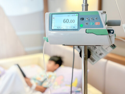 Medical infusion pump with digital screen displaying flow rate in a hospital room. Pediatric patient resting in bed in the background. Healthcare and intravenous therapy concept.