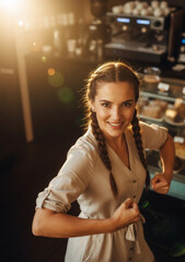 Fototapeta premium Confident female barista flexing her muscles in a coffee shop. Energetic young woman smiling at work. Small business and empowerment concept.