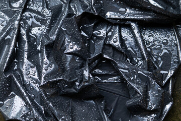 A dark, moody, abstract background of a crumpled black plastic trash bag with glistening water droplets from the rain