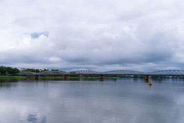 The historic Truong Tien Bridge spans the wide Perfume River under a dramatic, cloudy sky, its reflection subtly shimmering on the calm water's surface in Hue, Vietnam.