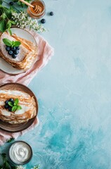 Overhead view of stacked crepes with blueberries and powdered sugar arranged on a textured blue surface with honey, cream, and floral accents