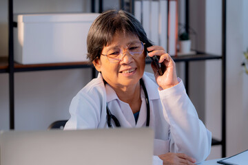 Senior asian doctor speak on smartphone while working on laptop in medical office, offering telemedicine consultation, healthcare and digital technology, elderly female physician online consultation.