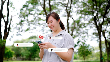 Cheerful Asian woman smiles while using a smartphone to read comments and interact with followers on a social media app, surrounded by like and message icons in a park.