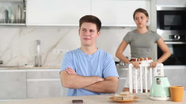 Annoyed young guy sitting at the table while his girlfriend is quarreling with him standing behind in the kitchen