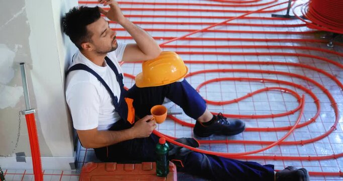 Tired construction worker resting on floor during underfloor heating installation, drinking coffee and showing fatigue and stress after hard work.