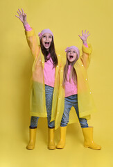 Two little sisters in yellow raincoats, boots and hats on a yellow background in autumn