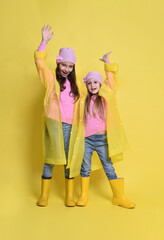Two little sisters in yellow raincoats, boots and hats on a yellow background in autumn