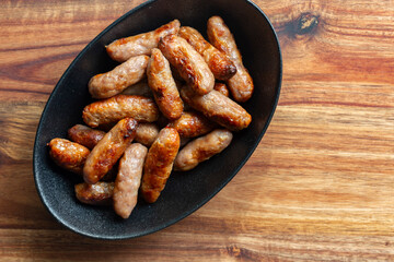 Cooked party cocktail sausages in a black stoneware dish background backdrop. Acacia wood chopping board background.