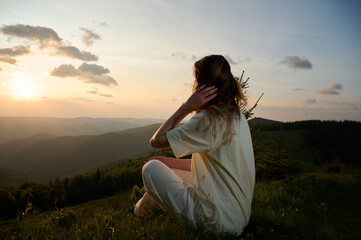 Woman sits peacefully on a grassy hill, gazing at the sunset as golden light bathes the landscape....