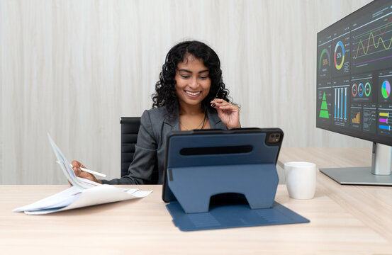 A businesswoman with curly hair smile as she examine data on her tablet while sitting at a desk in a sleek office environment. She has document and a coffee mug nearby.