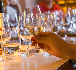 Close-up of a hand holding a glass of white wine during a wine tasting session. Transparent glass with golden liquid reflecting light, surrounded by other glasses on the table. Concept of luxury