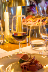 Close-up of a red wine glass on a dinner table with gourmet snacks, cheese, charcuterie, and appetizers. Warm evening atmosphere with elegant lighting, people in background, and luxury dining setting
