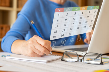 Businesswoman writing notes while using a digital calendar and laptop, managing her schedule, tasks, and reminders for efficient workflow and project planning