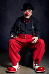 Clown on dark background. Dressed in red pants and a black shirt with suspenders, red clown nose and bowler hat on head.