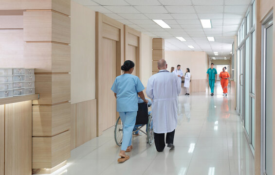 Two health care professional walk with a patient in a wheelchair down a well-lit hospital corridor, where multiple staff member are attending to their duty.