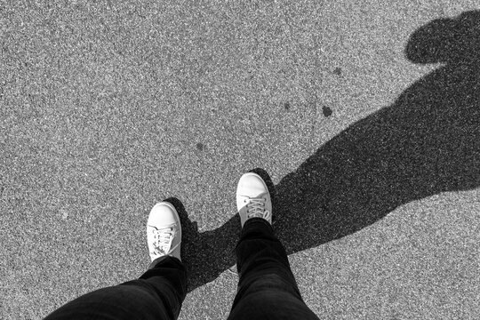 top view of a person wearing white sneakers standing on asphalt, with a long shadow cast on the ground