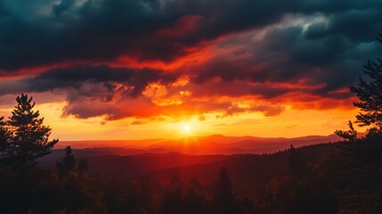 Dramatic sunset over a mountain range. Dark silhouettes of trees frame a vibrant fiery orange and red sky