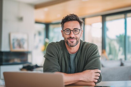 Smiling young businessman with glasses working on laptop at home office. Concept of remote work, freelancing, entrepreneurship, productivity, success, modern digital lifestyle. - Powered by Adobe