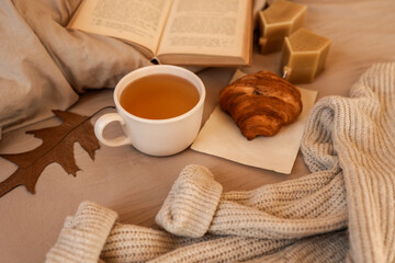Cozy autumn morning scene with tea, croissant, and a book on a bed.
