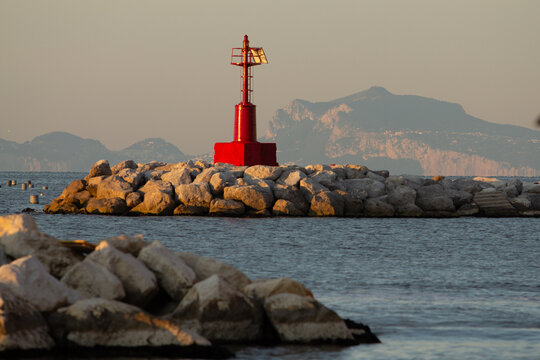 A striking red lighthouse stands on a rocky breakwater at sunset, with a misty mountain range in the background. Excellent for travel brochures, sailing magazines, and atmospheric coastal-themed conte - Powered by Adobe