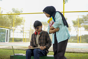 A dedicated female Muslim tutor wearing a hijab provides one-on-one mentorship to a young male student studying with a laptop outdoors