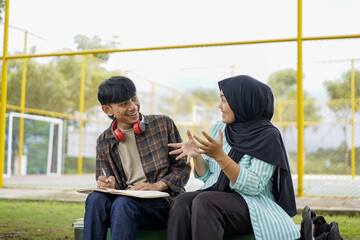 A cheerful young Asian man listens intently to his female Muslim friend in a hijab as they collaborate on a creative project outdoors on campus