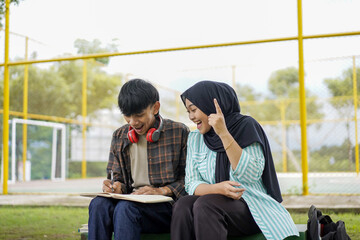 A moment of inspiration between two diverse Asian college students studying outdoors. A smiling young woman in a hijab explains her creative idea to a male classmate writing in a notebook