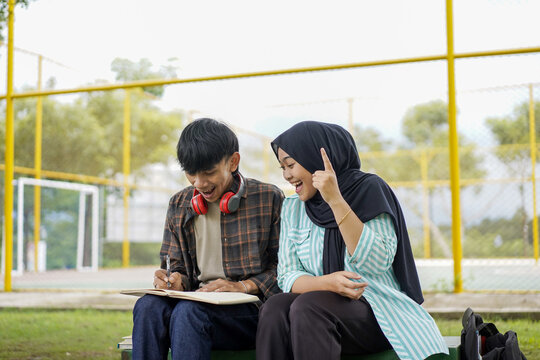 A moment of inspiration between two diverse Asian college students studying outdoors. A smiling young woman in a hijab explains her creative idea to a male classmate writing in a notebook