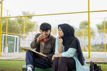 A happy young man and a Muslim woman in a hijab sitting together outdoors, collaborating on a school project and sharing ideas while looking at a digital tablet