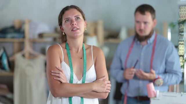 Upset young female dressmaker being scolded by angry man standing behind in sewing workshop. High quality 4k footage