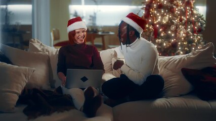 Joyful man and woman celebrating Christmas, using laptop at home and chatting. Portrait of biracial loving couple in cozy interior of living room, happy New Year fest, winter vacation, good mood
