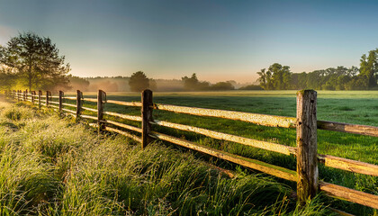 Obraz premium Sunlit Rural Landscape with Wooden Fence, Misty Field, and Trees at Sunrise morning