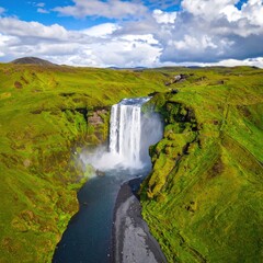 Obraz premium Aerial view of a waterfall crashing down into a rocky riverbed amidst vibrant green hills, under a partially cloudy sky