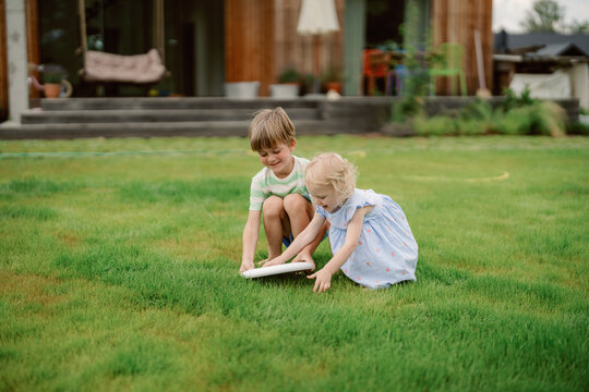 Two children enjoy a playful moment on lush grass in a backyard. The boy and girl interact joyfully while exploring a flat object during bright daylight. - Powered by Adobe