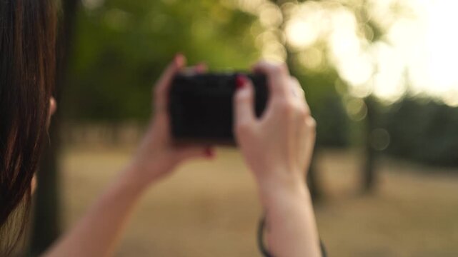 A young woman, closeup from behind, using a digital mirrorless camera and taking photos of landscapes in the park during the golden hour in the park
