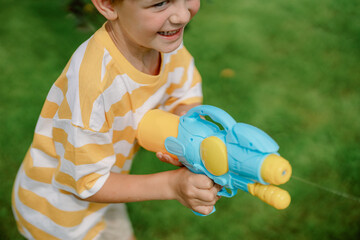 A joyful child wearing a striped yellow shirt is playing with a bright blue and yellow water gun in a green backyard. The weather is sunny, creating a fun atmosphere for outdoor play.
