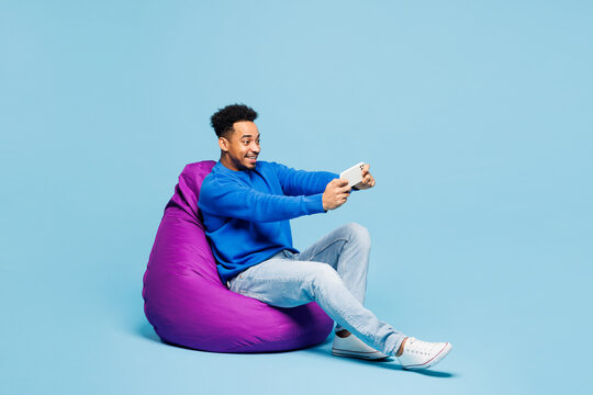 Full body young man of African American ethnicity wears sweatshirt clothes sit in bag chair use play racing app on mobile cell phone hold gadget for pc video games isolated on plain blue background. - Powered by Adobe