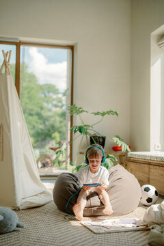 A young boy sits on a comfortable bean bag, wearing headphones and focused on a tablet, surrounded by a cozy living space with natural light and plants.