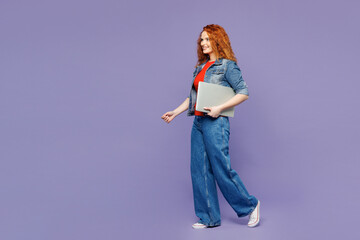 Full body side view young ginger woman she wearing red t-shirt denim shirt hold closed laptop pc computer walk go isolated on plain pastel light purple background studio portrait. Lifestyle concept.