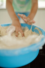 A child is using their hands to mix dough in a large blue bowl on a kitchen counter. Flour is visible, creating a playful and messy atmosphere. The light-filled kitchen enhances the joyful moment.