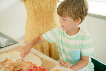 A young boy spreads sauce on pizza dough while a family member watches. The kitchen is well-lit, creating a cheerful atmosphere for cooking together.