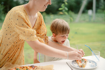 A mother and her child are having fun together outdoors. The child is excitedly reaching for a slice of pizza as the mother adds toppings, creating a joyful family moment on a sunny day.
