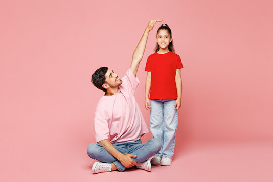 Full body young parent man with child girl wearing red clothes. Daddy little kid hold hand above head show how much she grown, try to be taller isolated on plain pink background. Father's Day concept. - Powered by Adobe