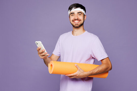 Young fitness trainer instructor sporty man sportsman wear headband t-shirt training in home gym hold yoga mat use mobile cell phone isolated on plain purple background. Workout sport fit abs concept.