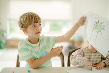 A young boy shows off his vibrant drawing in a cheerful classroom. Another child is seated nearby, engaging in a creative activity, surrounded by a bright and inspiring environment.