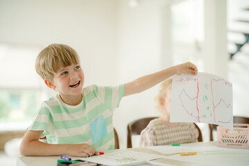 A young child proudly displays a colorful drawing while sitting at a table. The atmosphere is bright and cheerful, with another child engaged in activities nearby.