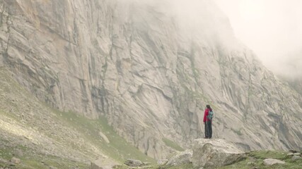 Indian woman traveler in red jacket with rucksack standing on a cliff in Hampta Pass, joyful and adventurous, showcasing trekking lifestyle.
